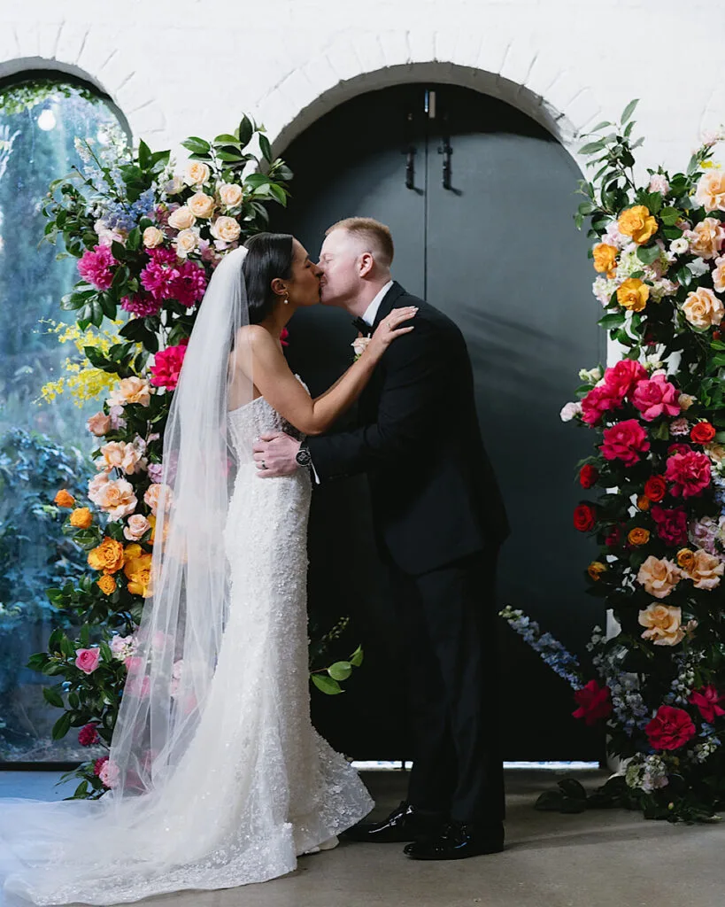 Couple sharing their first kiss beneath a vibrant Bloom Republica floral arch of roses, peonies and poppies