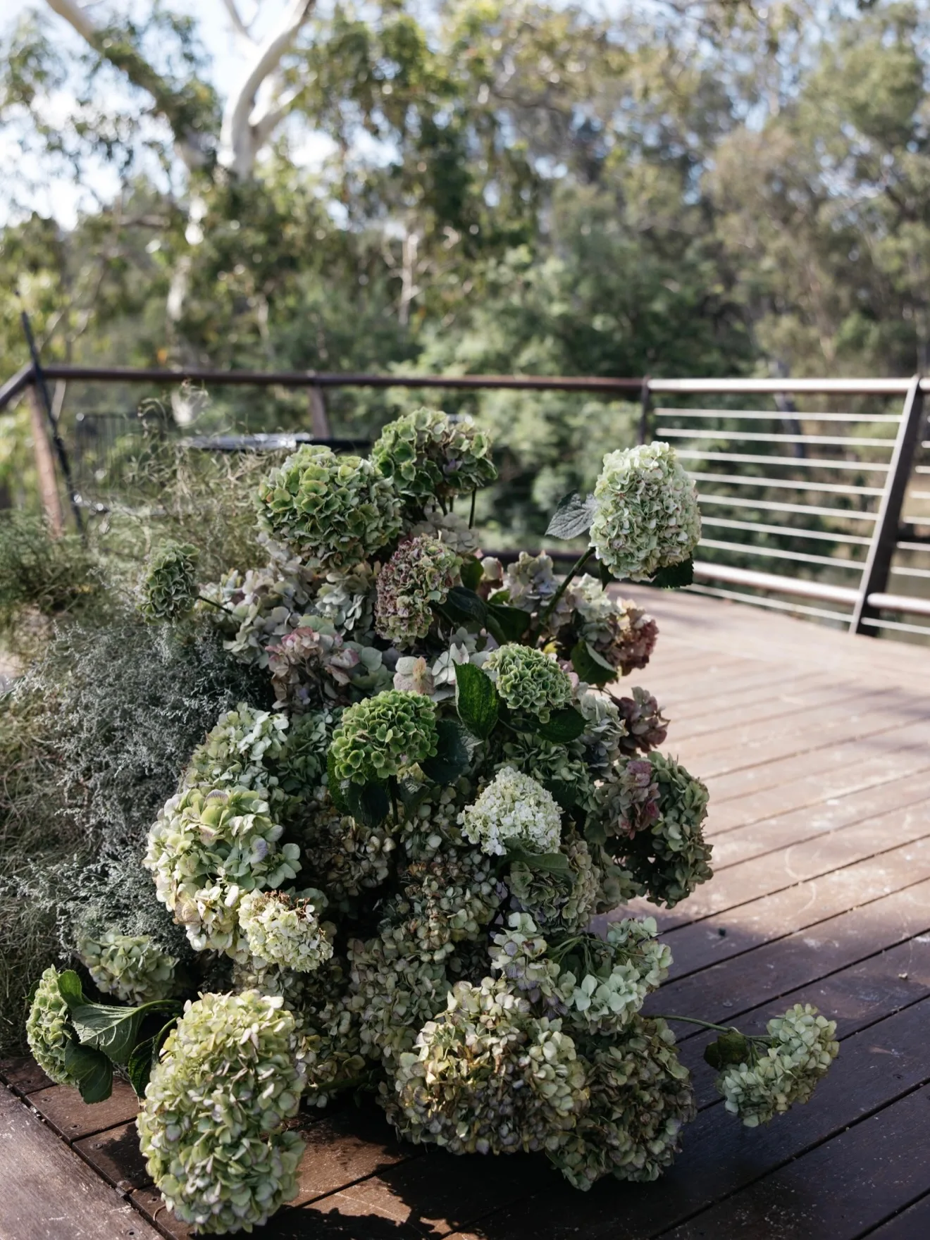 Antique-toned hydrangea and green foliage ground arrangement on a deck with bushland behind