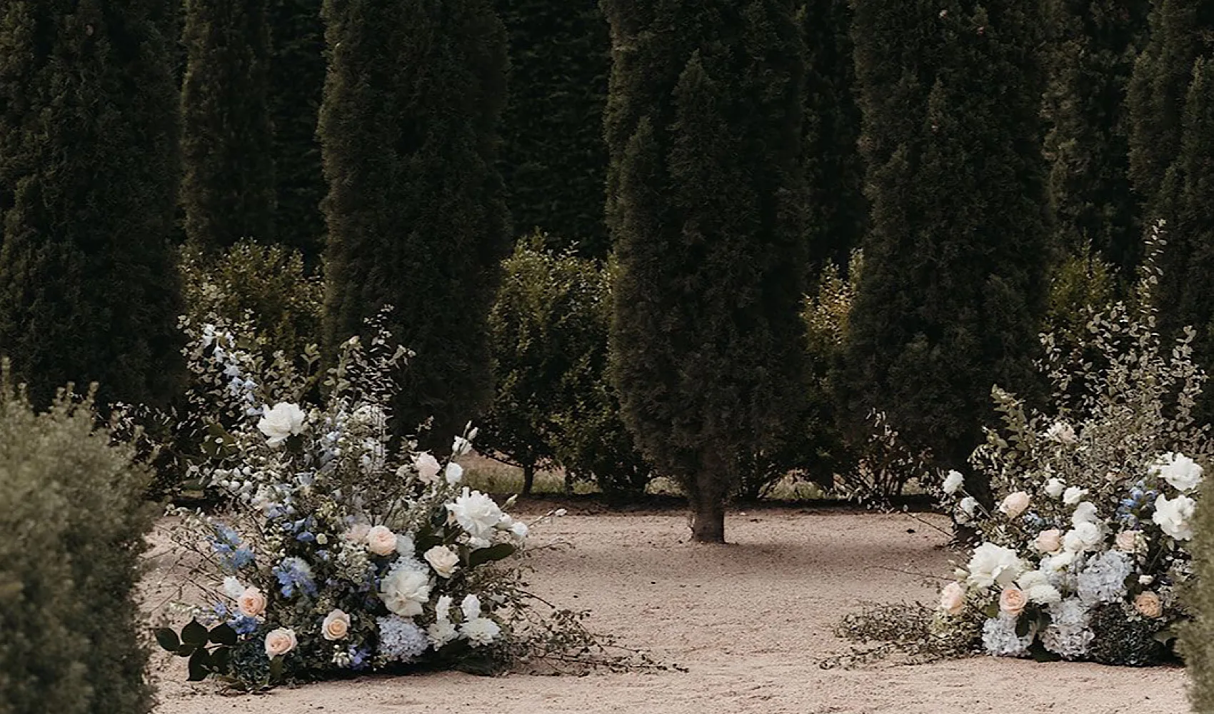 Two soft meadow arrangements flanking a cypress-lined outdoor ceremony aisle