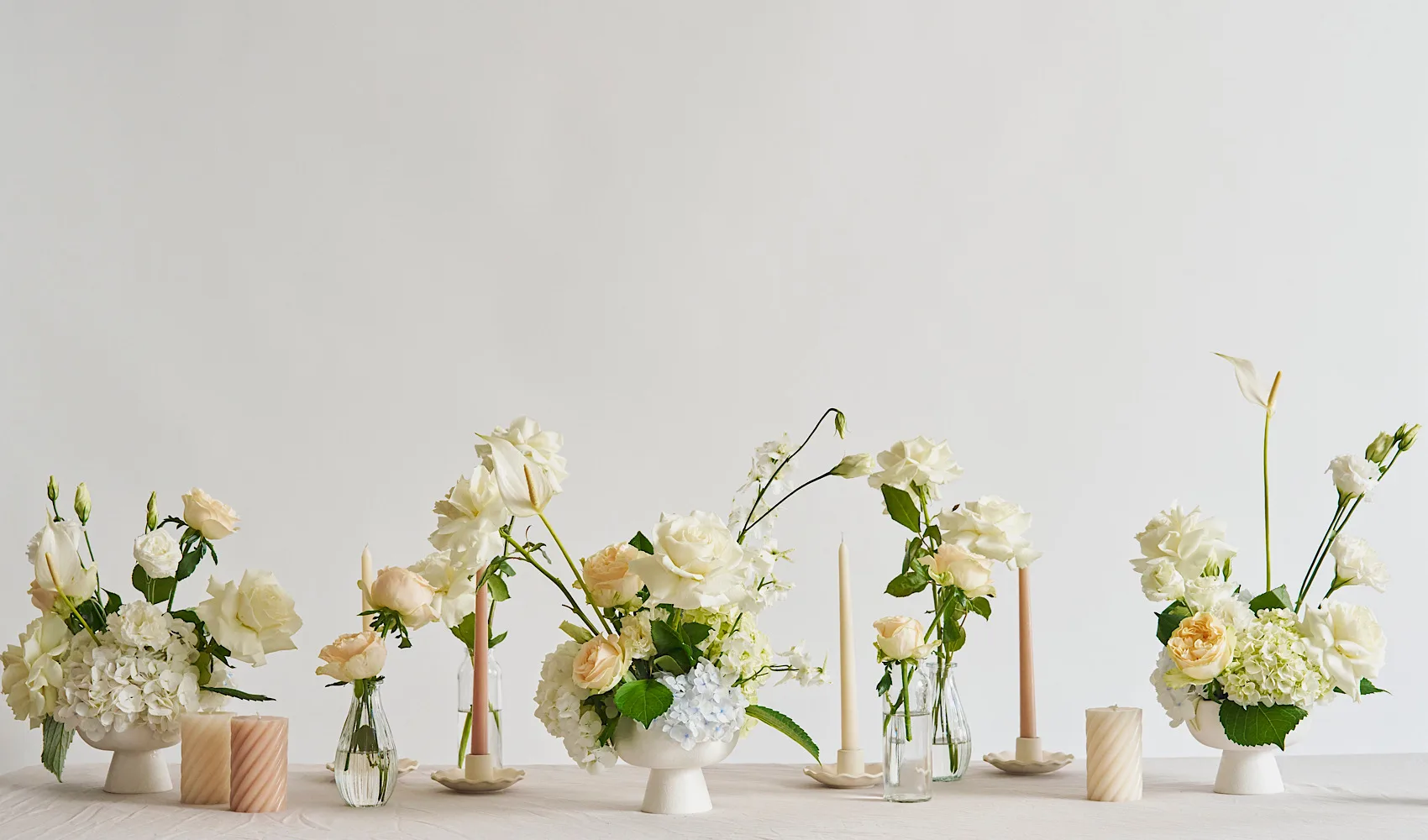 An editorial run of white and ivory Bloom Republica arrangements in footed bowls and bud vases, styled with taper candles along a pale table