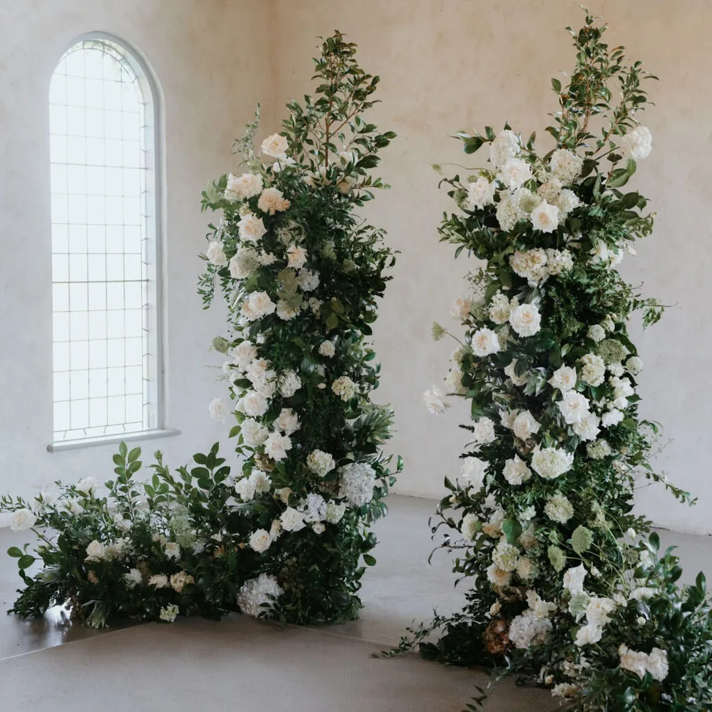 Two tall white floral ceremony pillars in a plaster room with an arched window
